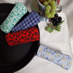 May include: A black cast iron frying pan with four fabric handle covers. The covers feature floral, paw print, and gingham patterns in blue, red, and teal. A small ceramic cup with grapes is in the background.