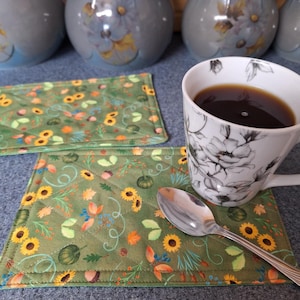 Peut inclure: Deux sets de table rectangulaires en tissu matelassé avec un fond vert orné d'un motif de tournesols, de petites citrouilles, de glands et de feuilles. Une tasse en céramique blanche à motif floral noir remplie de café et une cuillère en argent.