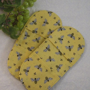 May include: Two yellow oven gloves with a bee pattern. The oven gloves are made of fabric and have a quilted design.