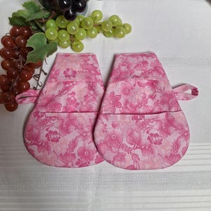 May include: A pair of pink floral oven mitts with a pocket on each mitt. The mitts are made of a soft, quilted fabric and are ideal for protecting your hands from heat.