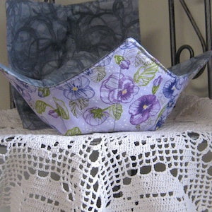Peut inclure: Un bol en tissu blanc avec un motif floral violet et vert repose sur un napperon en crochet blanc. Le bol est plié en deux et a une doublure en tissu gris.
