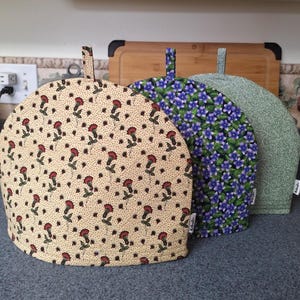 Peut inclure: Trois couvre-théières alignés, chacun avec un motif floral différent. Le premier est beige avec des fleurs rouges, le second a des fleurs bleues et le troisième est vert avec un motif subtil. Chacun a une poignée en boucle.
