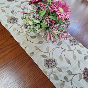 May include: A beige table runner featuring a floral design in brown and gold hues, complemented by a bouquet of pink and green flowers. The runner is displayed on a wooden table, serving as a decorative element.