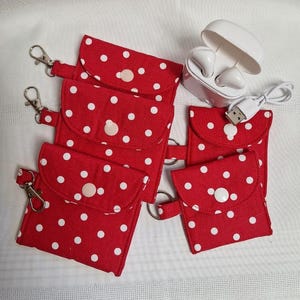 May include: Four red fabric pouches with white polka dots, each featuring a flap with a snap button closure and a metal swivel lobster clasp. A white wireless earbud charging case with an open lid and a white USB charging cable are included.