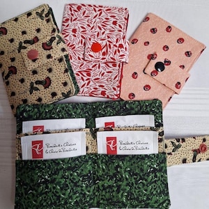 May include: Several fabric tea wallets with different patterns. One is green with a leaf design, another is tan with floral accents, a third is red with white floral, and a fourth is pink with ladybug prints. Each has a button closure.