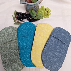 May include: Four oven mitts in green, teal, yellow, and blue, each with a white swirl pattern. The mitts are arranged in a row, with a jug of grapes and a bunch of grapes in the background. The mitts are made of fabric.