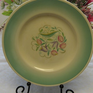 Peut inclure: Une assiette en céramique blanche avec un bord vert et un motif floral composé de fleurs roses, jaunes et vertes. L'assiette est sur une nappe blanche.