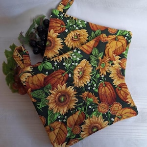 May include: Two square quilted pot holders featuring a dark green background with a repeating pattern of orange pumpkins, yellow sunflowers, and green leaves. Each pot holder includes a small fabric loop at the corner for convenient hanging storage.