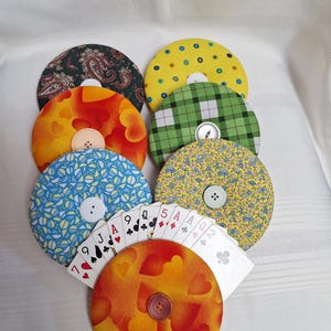 May include: A set of seven circular fabric-covered card holders, each featuring a central button. The patterns include paisley, yellow polka dots, green plaid, orange hearts, blue geometric shapes, and yellow floral prints, holding a fan of playing cards.
