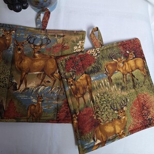 May include: Two brown and green pot holders with a deer and forest scene. The pot holders have a loop handle.