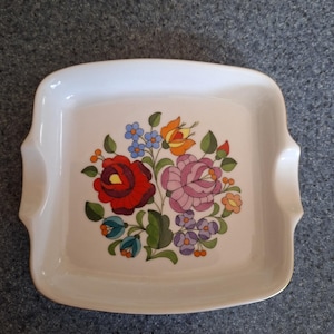 May include: A white, square ceramic serving dish with a floral design. The colourful floral pattern includes red, pink, orange, blue, and purple flowers with green leaves. The dish has two small handles.