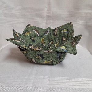 May include: A green fabric bowl with a fish pattern. The bowl is made of a textured fabric and has a unique, folded design. The fish are in various colours, including yellow, brown, and white.