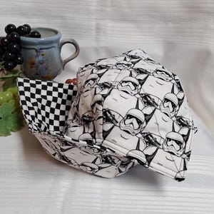 Peut inclure: Un bol en tissu noir et blanc avec un motif à carreaux à l'intérieur et un motif de visages de Stormtroopers à l'extérieur. Le bol est plié et posé sur une surface blanche.