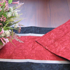 May include: A red and black table runner with a floral pattern. The runner is folded in half and has a white stripe down the centre. A bouquet of pink flowers is in the foreground.