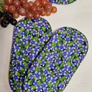 Peut inclure: Deux maniques en tissu de forme ovale avec un motif répétitif de petites fleurs violettes à cœur jaune et de feuilles vertes sur fond noir. Ces articles matelassés présentent des bords arrondis et un imprimé floral uniforme.