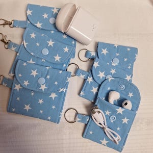 Peut inclure: Une collection de pochettes en tissu bleu clair avec un motif d'étoiles blanches. Chaque pochette possède un rabat avec une fermeture à bouton-pression et un anneau porte-clés en métal. Une pochette contient des écouteurs sans fil blancs.