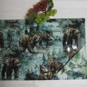 May include: A green and brown placemat with a pattern of bears and trees. A silver fork and spoon are tucked into a pocket on the right side of the placemat.