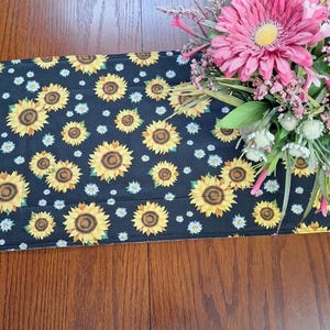 May include: A black table runner with a repeating pattern of yellow sunflowers and white daisies. The runner is on a wooden table.
