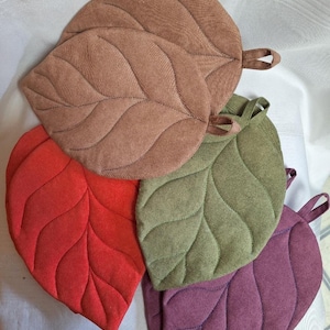 May include: A collection of four leaf-shaped pot holders in solid colours of brown, red, olive green, and purple. Each quilted fabric piece features stitched vein detailing and a small fabric loop at the top for hanging, arranged in a layered stack.