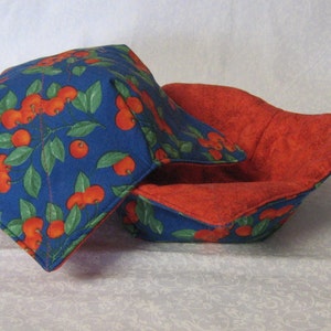 May include: Two quilted bowl cozies. The exterior features a blue fabric with a cherry branch pattern in red and green. The interior is a solid red fabric. The cozies are designed to hold bowls and protect hands from heat.