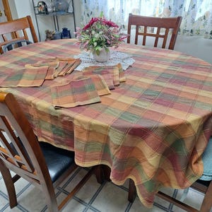 May include: A round dining table with a plaid tablecloth in shades of orange, green, and brown. The tablecloth has a subtle sheen and is draped over the table, with the edges hanging down. There are six chairs around the table, and a vase of flowers is in the centre.