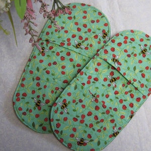May include: A pair of green oven mitts with a pattern of red strawberries and yellow bees. The mitts are oval shaped and have a seam down the middle.
