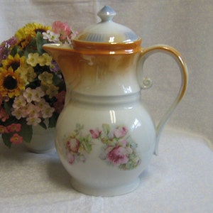 May include: A white porcelain teapot with a brown and blue lid and floral design. The teapot has a handle and a spout. The teapot is sitting on a white tablecloth.