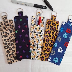 May include: Five fabric keychains with different patterns. The keychains are black with colourful hearts, white with grey paw prints, brown with orange leopard print, blue with red paw prints, and brown with tan leopard print.