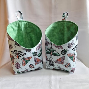 Peut inclure: Deux paniers de rangement suspendus en tissu vert et blanc avec un motif de papillons de nuit et de scarabées. Les paniers ont une doublure verte et un extérieur blanc avec un motif de papillons de nuit, de scarabées et de papillons.