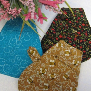 Peut inclure: Trois maniques matelassées aux motifs variés. L'une est turquoise avec un motif de tourbillon blanc, une autre est noire avec des fleurs rouges et blanches, et la troisième est beige avec des abeilles jaunes et des volutes blanches.