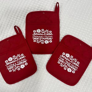 May include: Three red quilted oven mitts and pot holders with white floral designs and text. The text on each reads "Kathy's Kitchen", "Grammy's Kitchen", and "Winge Family Kitchen". Each has a loop for hanging.