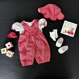 May include: A red and white gingham overall dress with a white blouse, a red and white gingham sun hat, a pair of white socks, a pair of white shoes, and a children's book titled "Bitty Bear at Play".