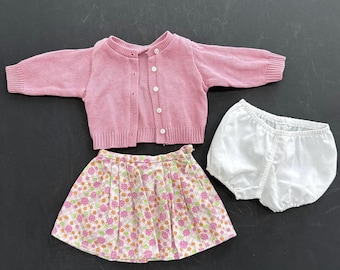 May include: A three-piece baby clothing set. The set includes a light pink button-up cardigan, a floral print skirt with pink, orange, and green flowers, and white bloomers. The clothing is made of soft fabric.