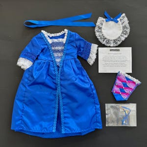 Könnte beinhalten: Ein blaues Puppenkleid mit weißer Spitzenborte, ein passendes blaues Band, ein weißer Spitzenkragen mit blauer Schleife, ein dekorativer Brustfleck mit rosa und blauen Bändern sowie eine Infokarte mit dem Titel American Girl Felicity's Stomacher.