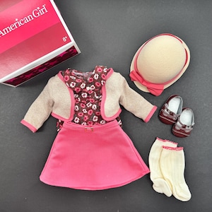 May include: A doll's outfit comprising a pink skirt, floral top, beige bolero jacket, matching hat with a pink bow, burgundy shoes, and cream socks. The box in the image reads "American Girl".