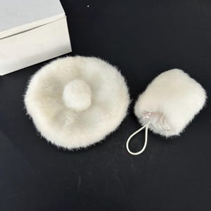 May include: A white, fluffy powder puff and a matching beret with a pom-pom. The beret is round with a small, central pom-pom. The powder puff is cylindrical with a drawstring. Both items are made of a soft, fur-like material.