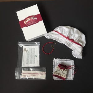 Puede incluir: Una caja blanca con la etiqueta The American Girls Collection, un gorro blanco con cinta roja, un collar de cuentas rojas, un paño cuadrado pequeño con borde de encaje, una bolsa estampada con cordón rojo y dos bolsas de plástico.