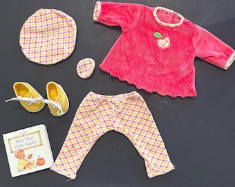 Puede incluir: Una camisa de terciopelo rosa con un bordado de manzana verde, pantalones de cuadros amarillos y rosas, un sombrero de cuadros amarillos y rosas y un libro titulado "Bitty Bear Picks Apples".
