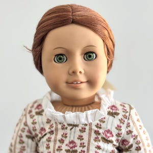 Puede incluir: Muñeca con cabello rojo y ojos verdes, vestida con un vestido con estampado floral y un cuello blanco con volantes. La muñeca tiene una expresión sonriente y mira a la cámara.