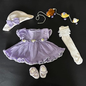 May include: A lavender dress with white lace trim, a white hat with a bow, white socks with lace trim, and white shoes. There are also three small toy animals.