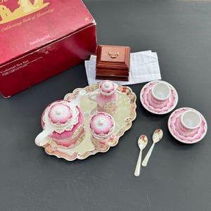 Könnte beinhalten: Ein Miniatur-Teeservice mit weißer Teekanne, Zuckerdose, Milchkännchen und zwei Tassen mit Untertassen, verziert mit einem rosa Blumenmuster. Das Set enthält zwei silberfarbene Löffel, ein silberfarbenes ovales Tablett, eine Holzschatulle und eine rote Schachtel.