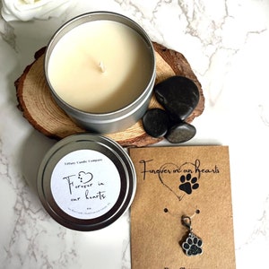 May include: A silver candle tin with a white label that reads "Forever in our hearts". The candle is surrounded by black stones. A brown card with the text "Forever in our hearts" and a paw print charm is also included.