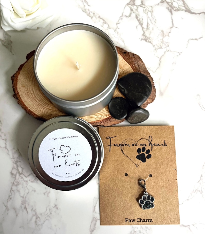 May include: A silver candle tin with a white label that reads "Forever in our hearts". The candle is surrounded by black stones. A brown card with the text "Forever in our hearts" and a paw print charm is also included.