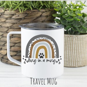 May include: White travel mug with a rainbow design and a paw print in the center. The text "hug in a mug" is written below the rainbow. The mug is sitting on a white surface.
