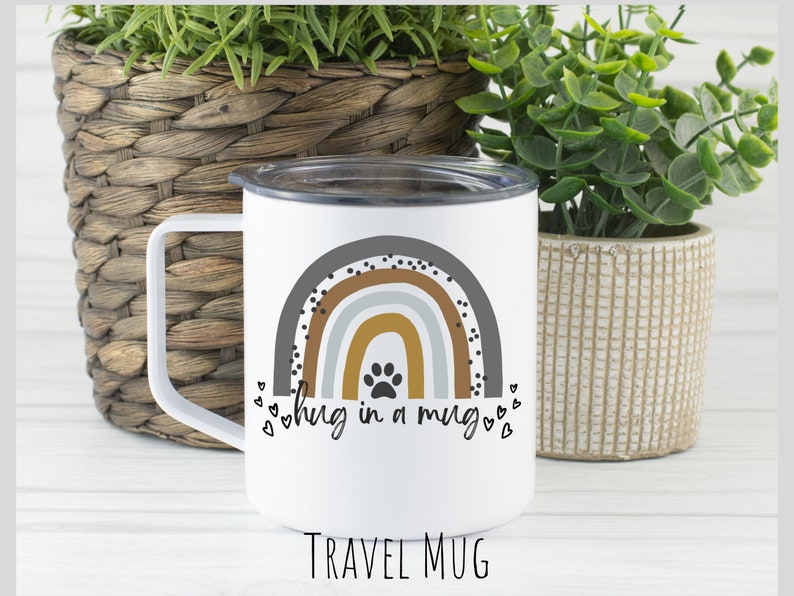 May include: White travel mug with a rainbow design and a paw print in the center. The text "hug in a mug" is written below the rainbow. The mug is sitting on a white surface.