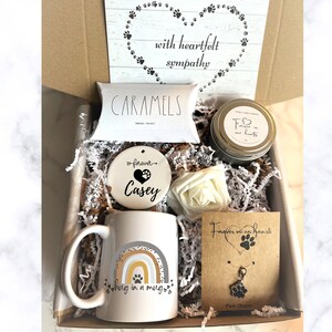 May include: A gift box filled with sympathy gifts for pet loss. The box contains a mug with a rainbow and paw print design, a ceramic ornament with the text "forever Casey", a candle with the text "Forever in our hearts", a bag of caramels, a white flower, and a necklace with a paw print charm.