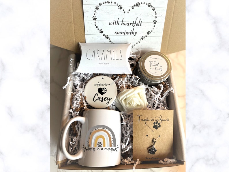 May include: A gift box filled with sympathy gifts for pet loss. The box contains a mug with a rainbow and paw print design, a ceramic ornament with the text "forever Casey", a candle with the text "Forever in our hearts", a bag of caramels, a white flower, and a necklace with a paw print charm.