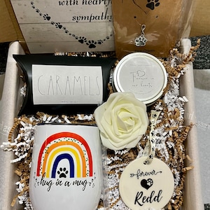 May include: A gift box filled with sympathy gifts for pet loss. The box contains a rainbow mug with the text "hug in a mug", a white rose, a candle with the text "Forever in our hearts", a small bag of caramels, and a ceramic ornament with the text "forever Redd".