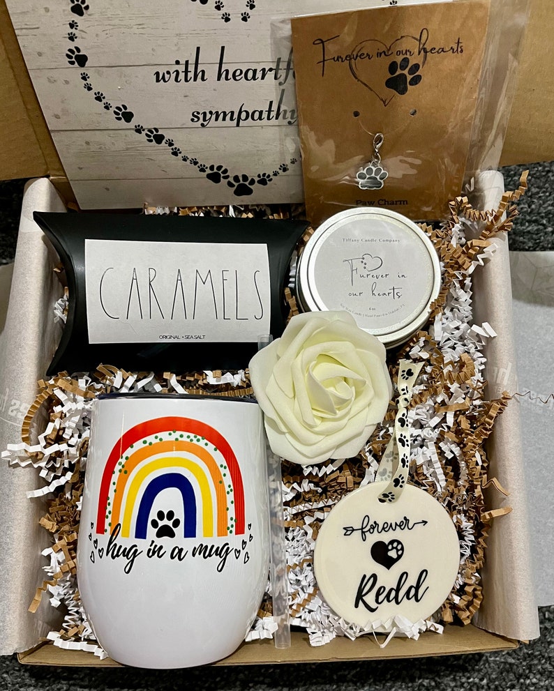May include: A gift box filled with sympathy gifts for pet loss. The box contains a rainbow mug with the text "hug in a mug", a white rose, a candle with the text "Forever in our hearts", a small bag of caramels, and a ceramic ornament with the text "forever Redd".