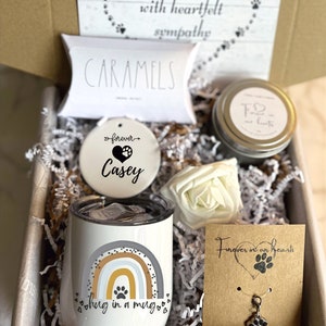May include: A sympathy gift box with a white ceramic ornament that says "forever Casey", a white ceramic mug with a rainbow and paw print design that says "hug in a mug", a small white box with the word "caramels" on it, a candle with a paw print and the words "Forever in our hearts", and a silver paw print charm on a brown card that says "Forever in our hearts" and "Paw Charm".